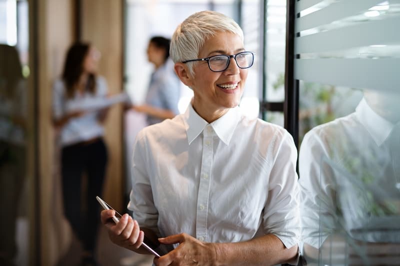 Senior female office manager standing near a corporate office's window, smiling while looking outside