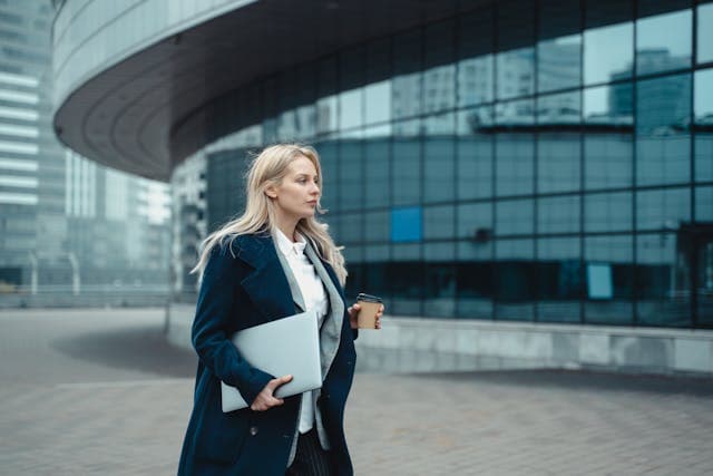 A business woman walking outside a modern building with a coffee cup on one hand and laptop in the other